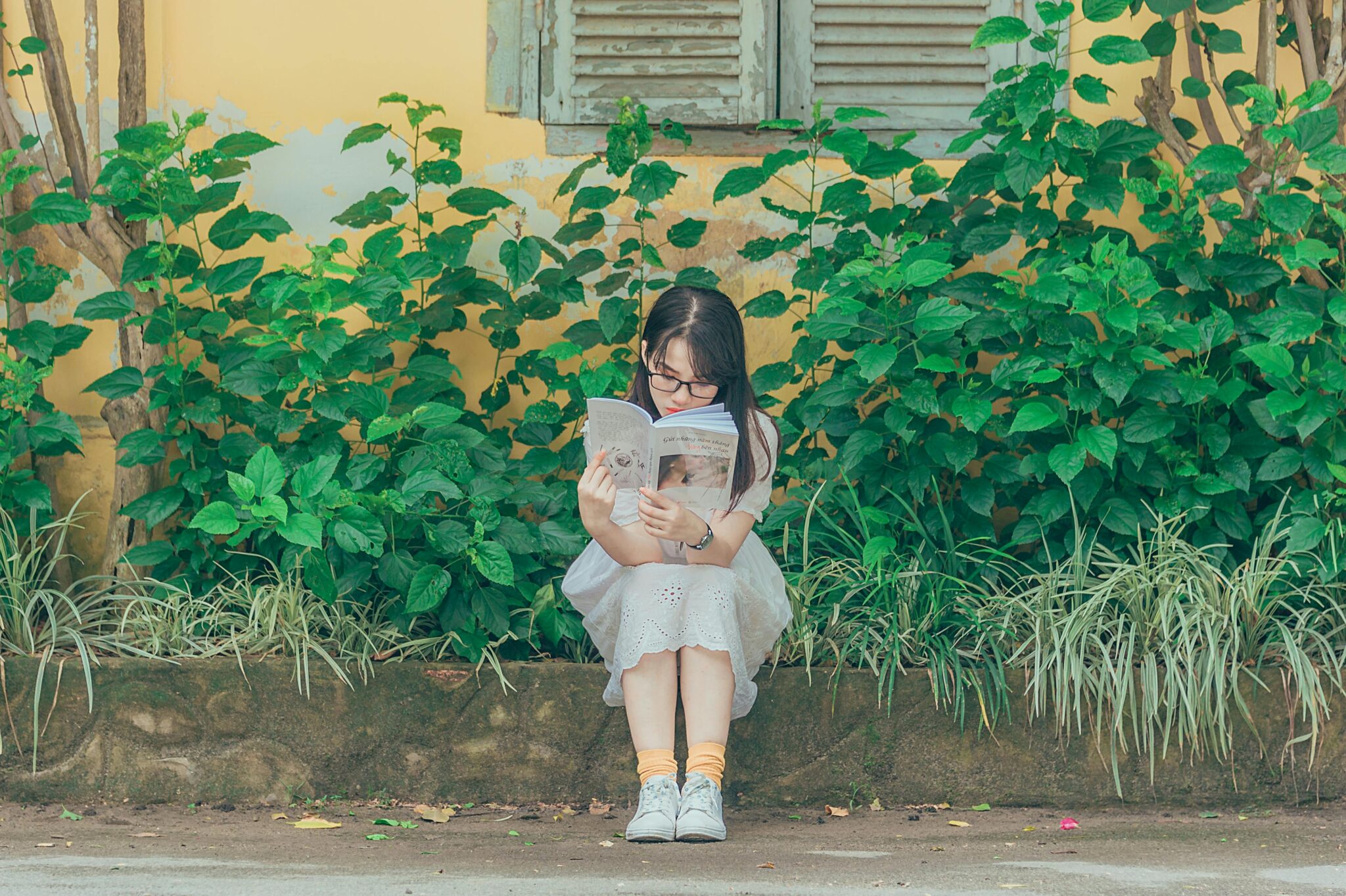 A young girl sits on a bench outdoors, immersed in a book with lush greenery around her.
