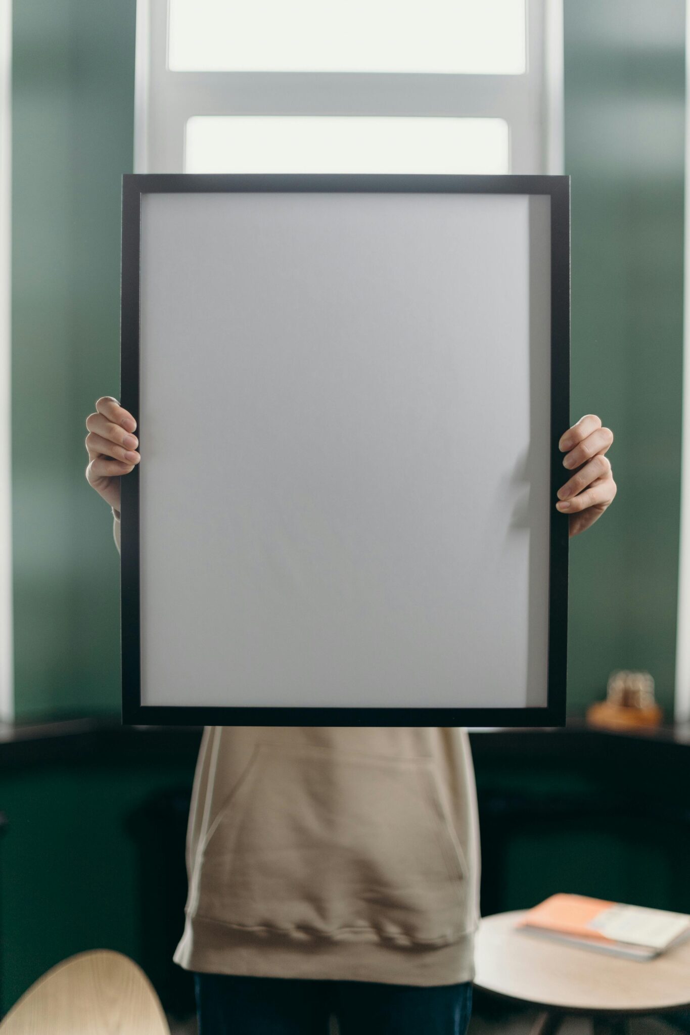 Anonymous person holds a blank vertical picture frame indoors against a window.
