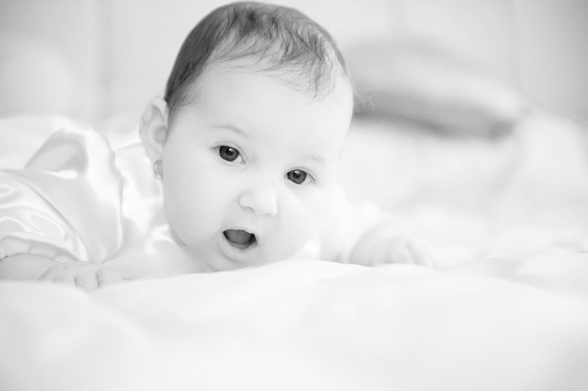 A cute black and white image of an adorable infant lying on a bed indoors.
